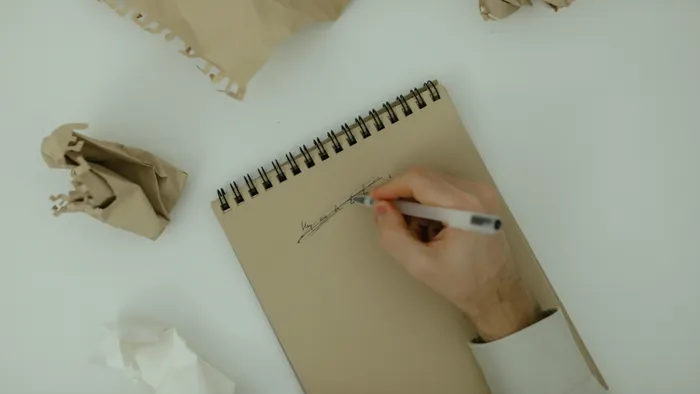 Mano escribiendo en una libreta rodeada de papeles arrugados sobre una mesa clara