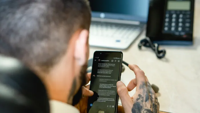 Persona leyendo una conversación larga en su teléfono, sentada en un escritorio con laptop y teléfono fijo